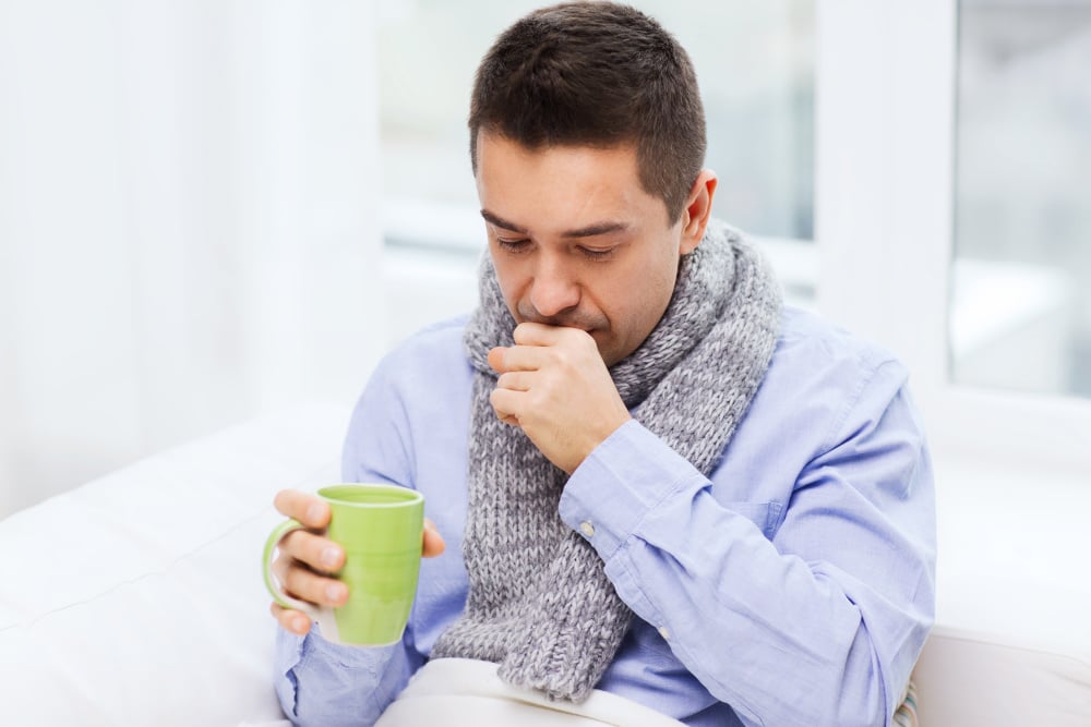 Sick man with a scarf around his neck sitting on a couch and holding a hot beverage with his hand